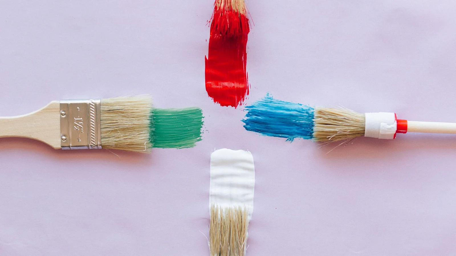 Paintbrushes dipped in red, blue, green and white paint arranged in a cross on a pale pink surface