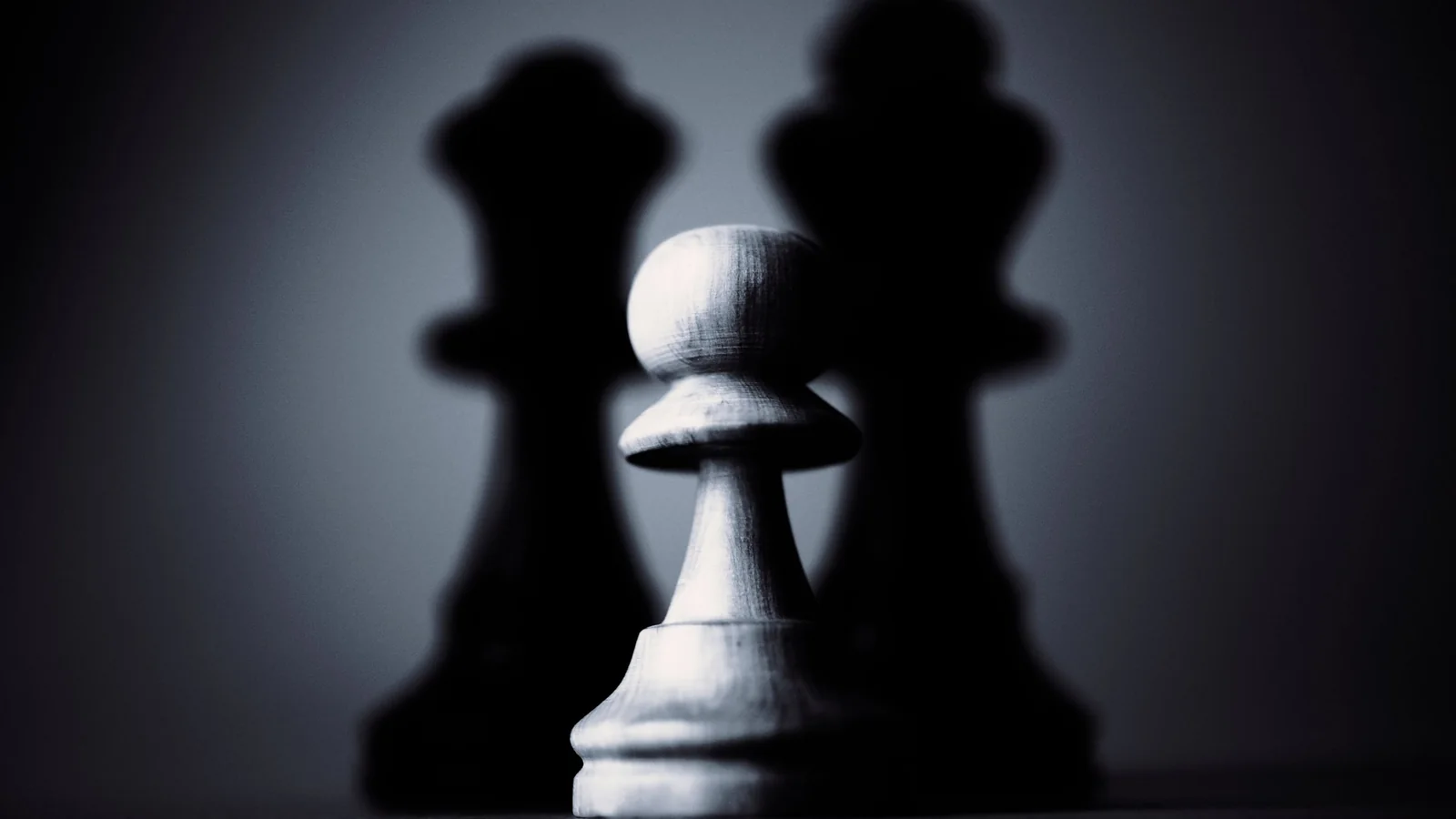 A white chess pawn lit from the side, casting tall dark shadows of chess pieces on the wall behind it